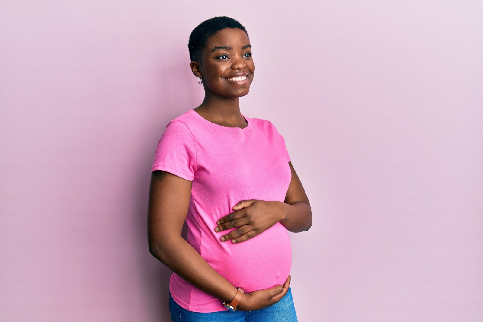 Young african american woman expecting a baby, touching pregnant belly looking away to side with smile on face, natural expression. laughing confident